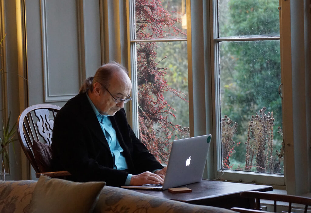 Balding Man with ponytail works on apple laptop by a window